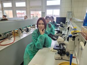  Uczniowie w zielonych fartuchach  laboratoryjnych siedzą przy stole laboratoryjnym, na którym stoją mikroskopy.